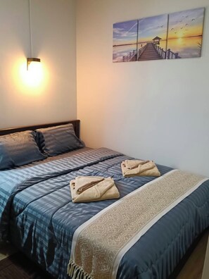 A bedroom with a bed, pillows, a blanket, a wall-mounted light, and a sunset painting. - SONGTHAI Muay Thai (Muang Krabi)