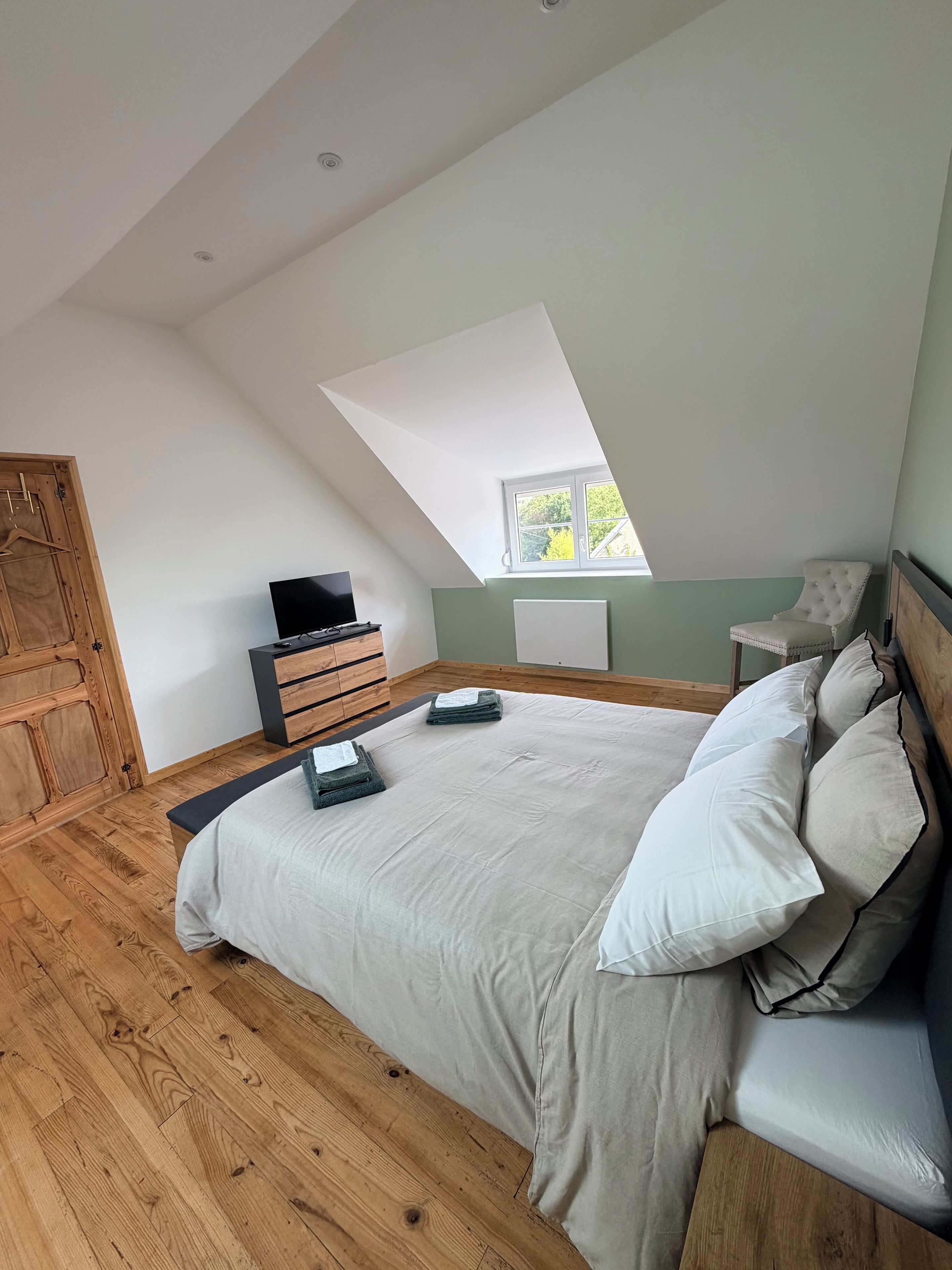 A bedroom with a sloped ceiling, a bed with pillows, a dresser, a television, and a window.
