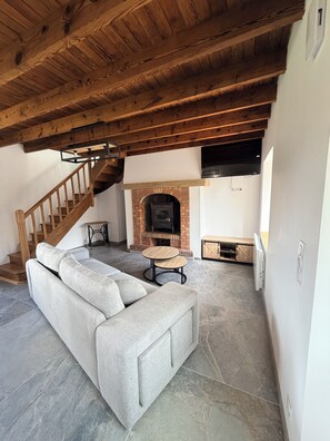 Un salon avec un sol en pierre, un canapé gris, un escalier en bois et une cheminée en briques.
