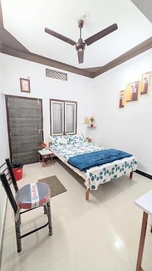 A bedroom with a bed, a chair, a desk, a ceiling fan, and a door. - Krishnaalay Guest House (Ujjain)