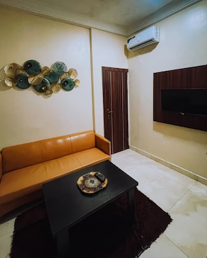 A living room with a sofa, a coffee table, decorative wall art, a door, and a mounted TV. - Alpines Hotels and Reservations (Port Harcourt)