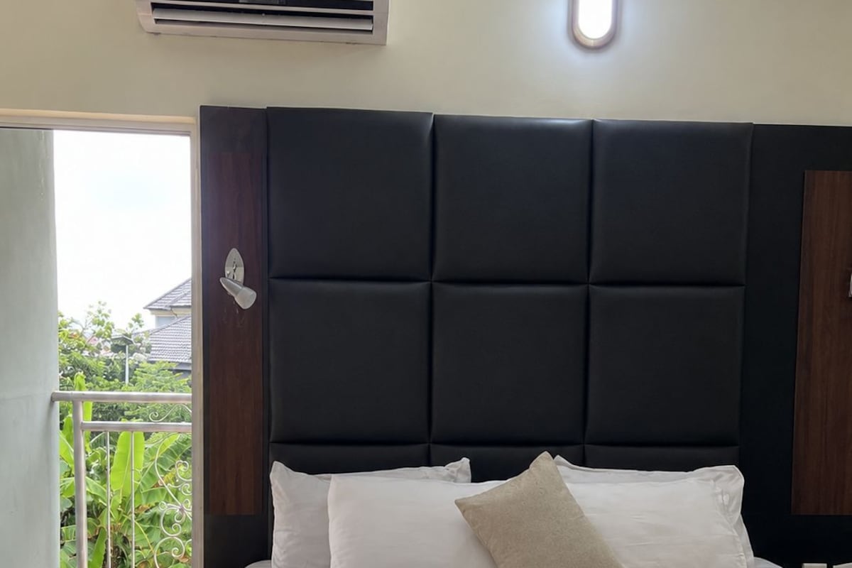A hotel room with a bed, a headboard, an air conditioning unit, a window, and a balcony with greenery.