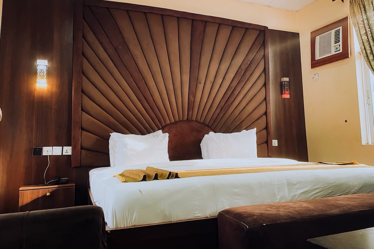 A hotel room with a large bed, a wooden headboard, a bedside table, and a wall-mounted air conditioning unit.