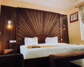 A hotel room with a large bed, a wooden headboard, a bedside table, and a wall-mounted air conditioning unit. - Alpines Hotels and Reservations (Port Harcourt)