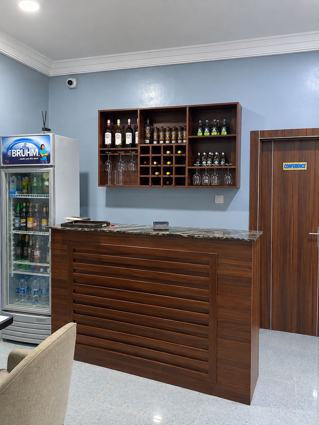 Un área de bar con mostrador de madera, un refrigerador lleno de bebidas y un cartel que indica una sala de conferencias.