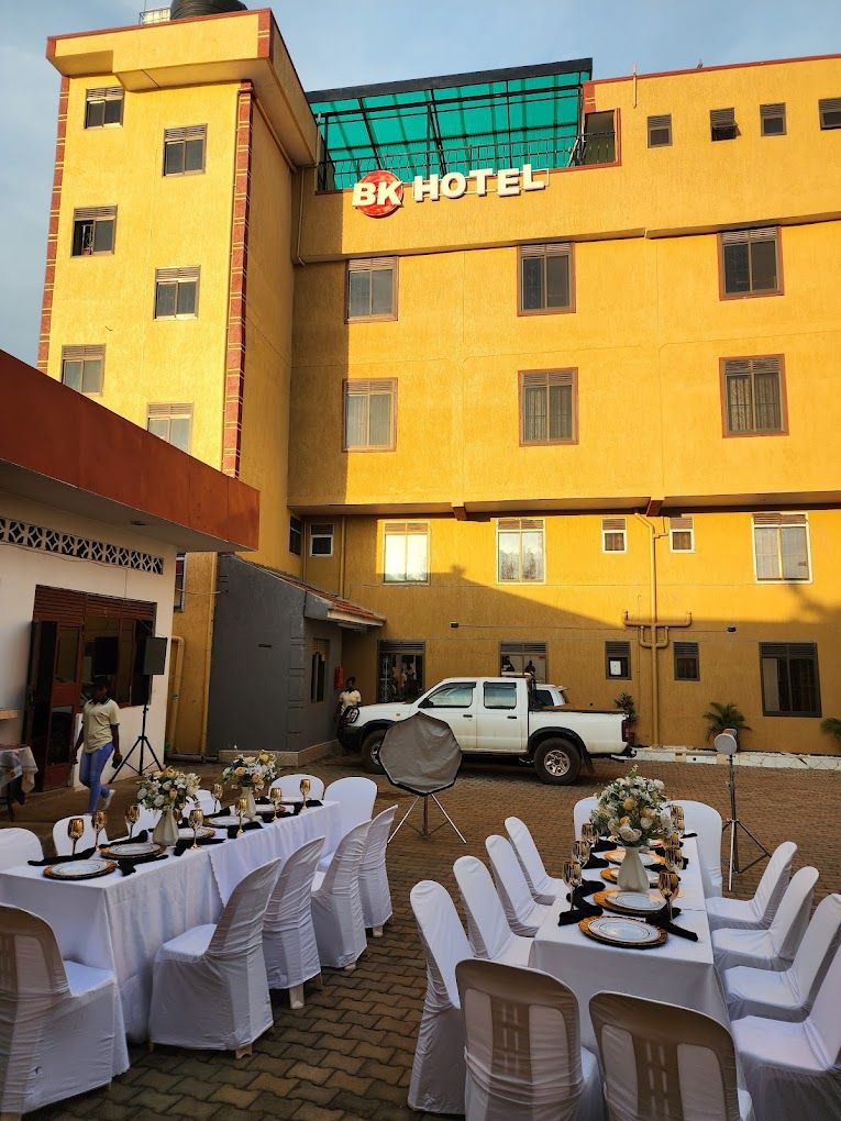Un hotel con fachada amarilla y un letrero que dice 'BK HOTEL'. Un camión pickup blanco está estacionado al frente, y hay mesas dispuestas para comer con manteles y sillas blancos.