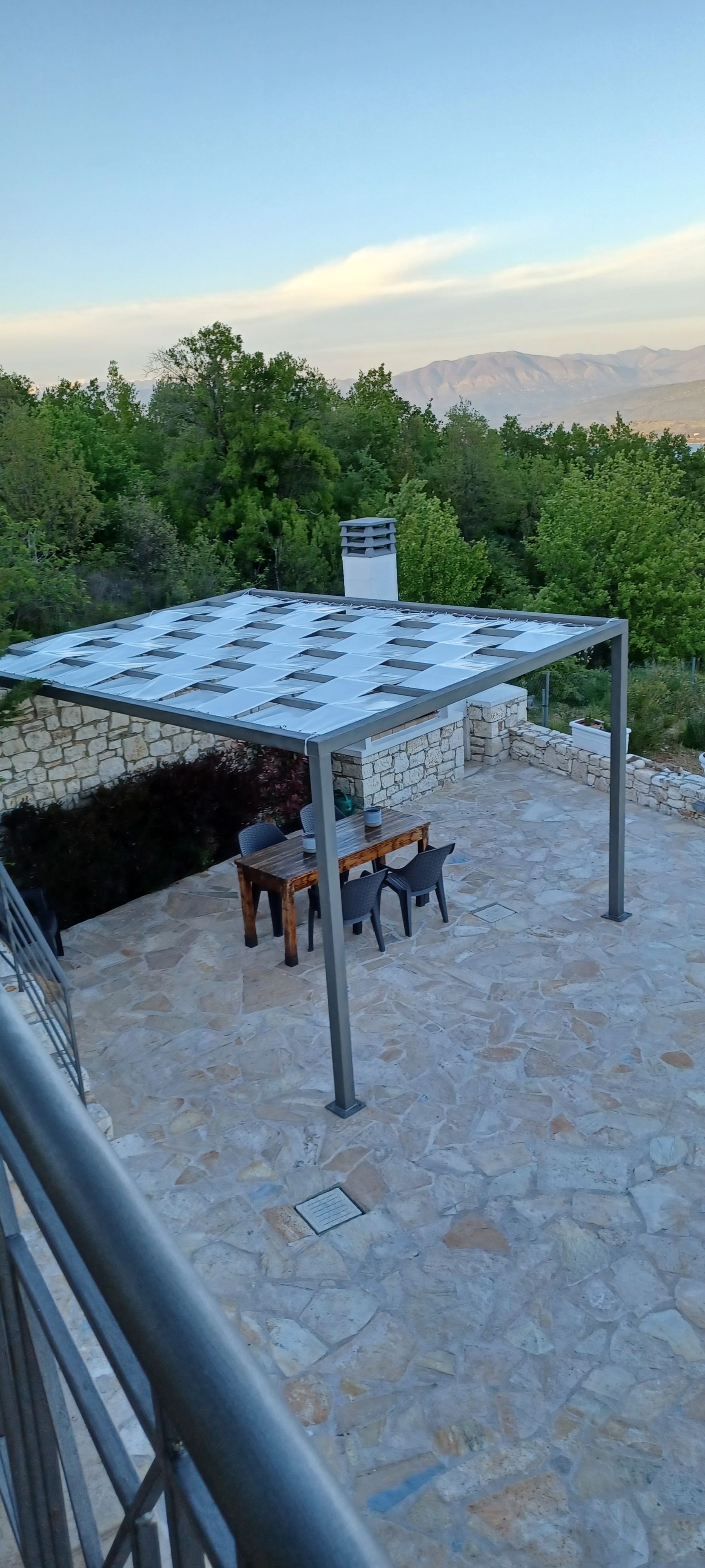 En stenbelagd uteplats med ett bord och stolar, en pergola med tak och en stenmur.