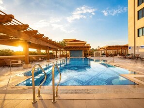 Una piscina con una pérgola y sillones de descanso, rodeada por un edificio con techo de tejas.