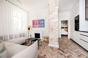 A modern living room with a sofa, a small table, and a fireplace. There is a brick accent wall and a kitchen area with cabinets and a refrigerator. - Wagner 12 Old Town Apartment - 2 bedrooms, sleeps 5, Wi-Fi (Rīga)
