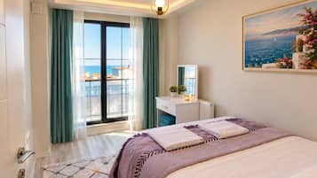 A hotel room with a bed, a desk, a chair, a window with curtains, a painting, and a view of the sea.
