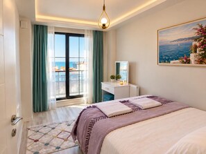 Habitación de hotel con cama, escritorio, silla, ventana con cortinas, un cuadro y vista al mar.