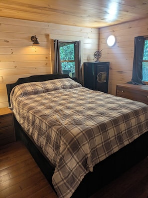 Une chambre dans une cabane en bois, comprenant un lit, une table de chevet, une commode, un radiateur et une fenêtre avec des rideaux.