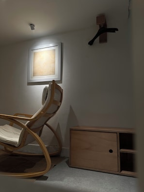 Una habitación con una silla de madera, un cuadro enmarcado en la pared, un armario pequeño y una lámpara en el techo.
