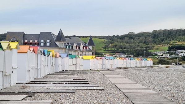 En række farverige strandhytter med en træbro, der fører ned til en stenstrand, foran huse og bakker.