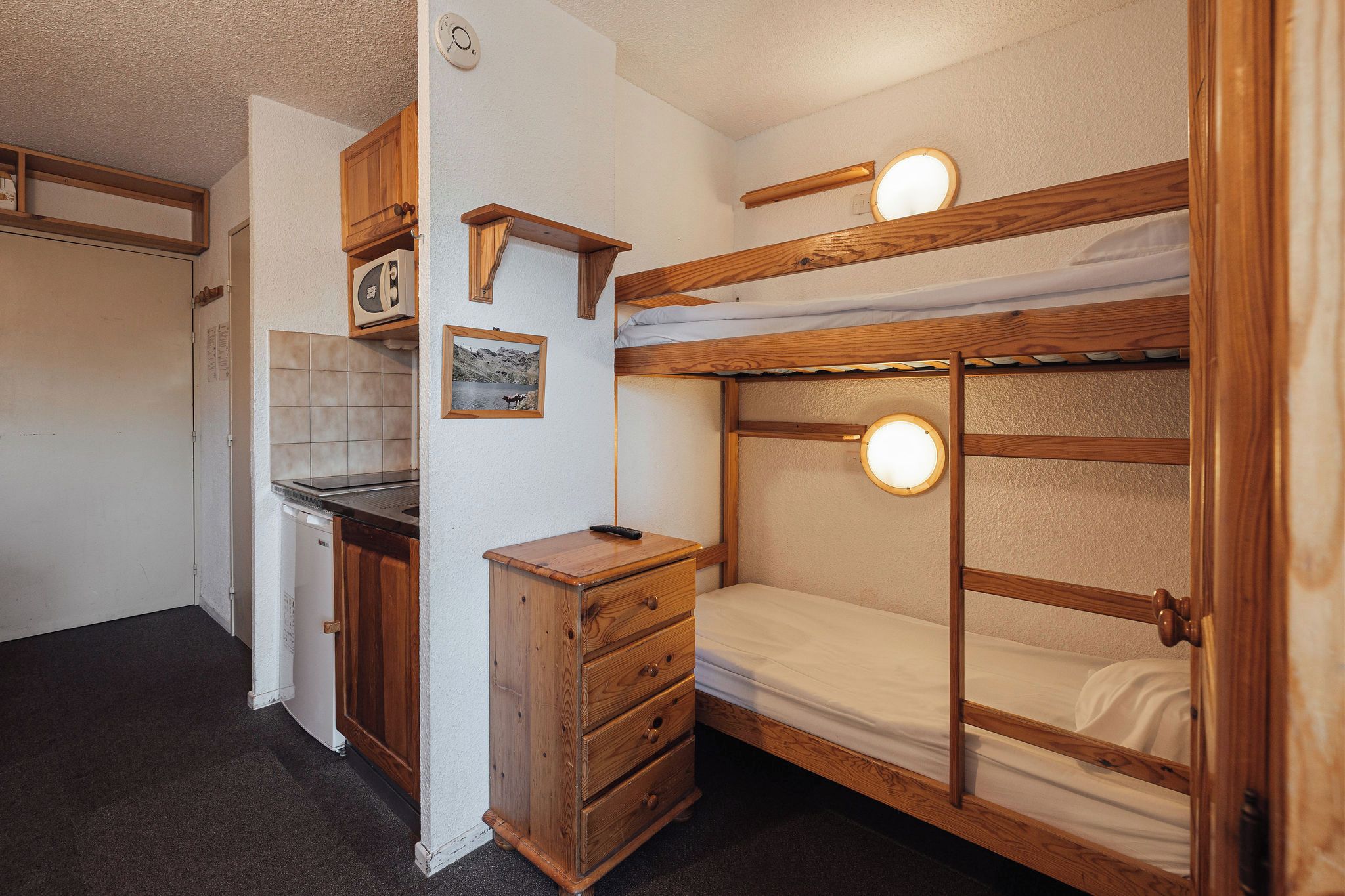 Een stapelbed met houten frame, een klein houten dressoir, een keuken met fornuis, en een wandlamp.