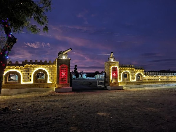 A brick gate with arched windows and a red door, illuminated by lights at dusk.