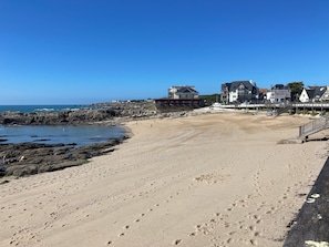 Une plage parsemée d'empreintes, un rivage rocheux et des bâtiments en arrière-plan.