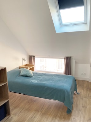 Une chambre avec un puits de lumière, un lit recouvert d’une courtepointe bleue, un plancher en bois et une table de chevet.