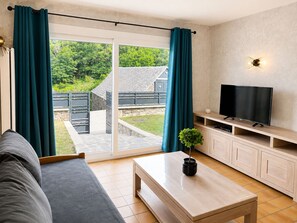 Una sala de estar moderna con un sofá, una mesa de centro, un televisor sobre un mueble de madera y una puerta corrediza de vidrio que da a un jardín.