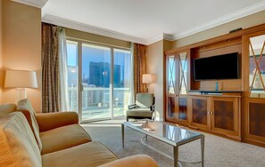 Habitación de hotel con sofá, sillón, televisión y vista panorámica de la ciudad a través de amplias ventanas.