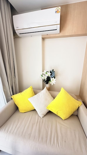 Een kamer met een beige bank, twee gele kussens, een wit kussen en een luchtverkoeler aan de muur.
