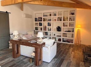 A room with a wooden ceiling, a white sofa, a wooden table with lamps, and a bookshelf filled with various items. - New Listing (Gujan-Mestras)