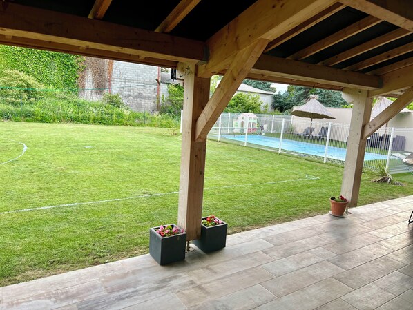 Un patio techado con vistas a un jardín trasero que incluye una piscina y un área de juegos cercada.