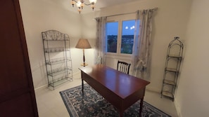 Quarto com mesa de madeira, luminária, cadeira, janela com cortinas e estante de metal.