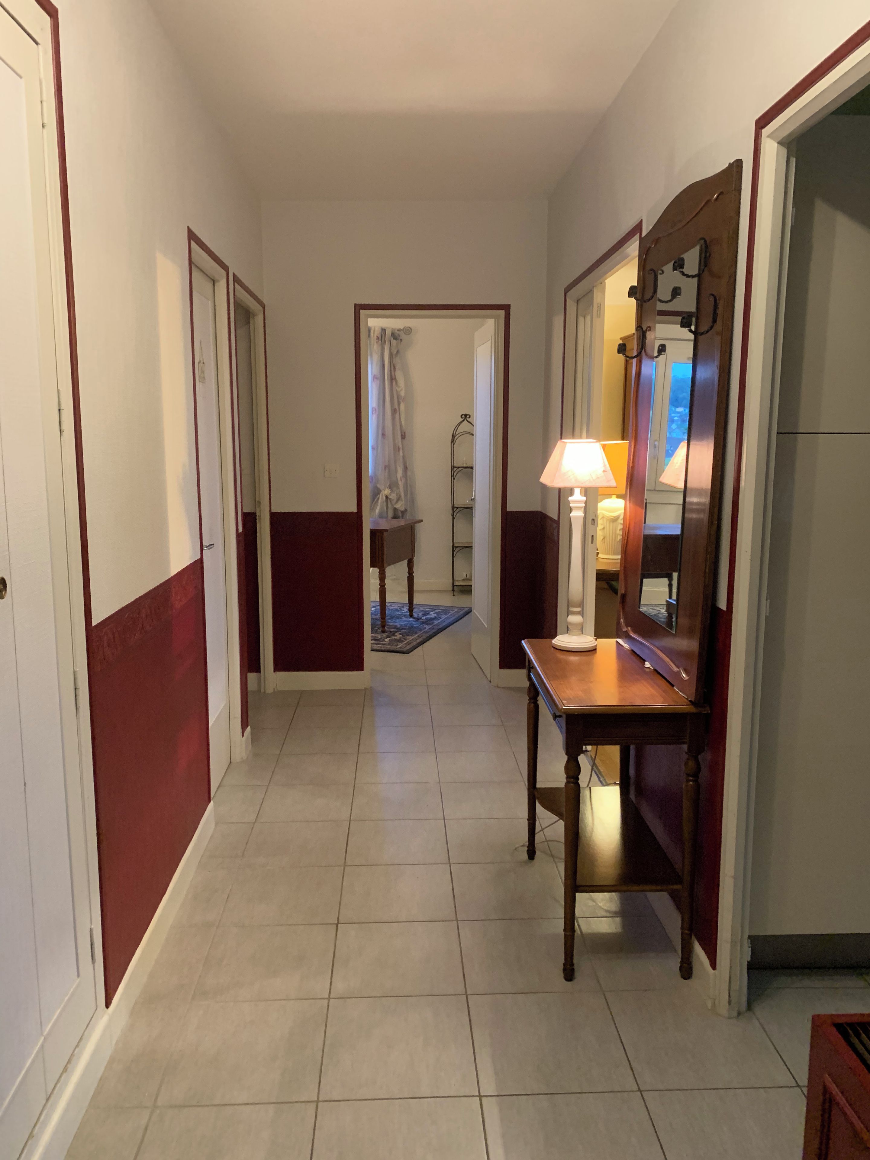 Un couloir avec une table en bois, une lampe et un miroir. Il y a une porte à gauche et une fenêtre à droite.