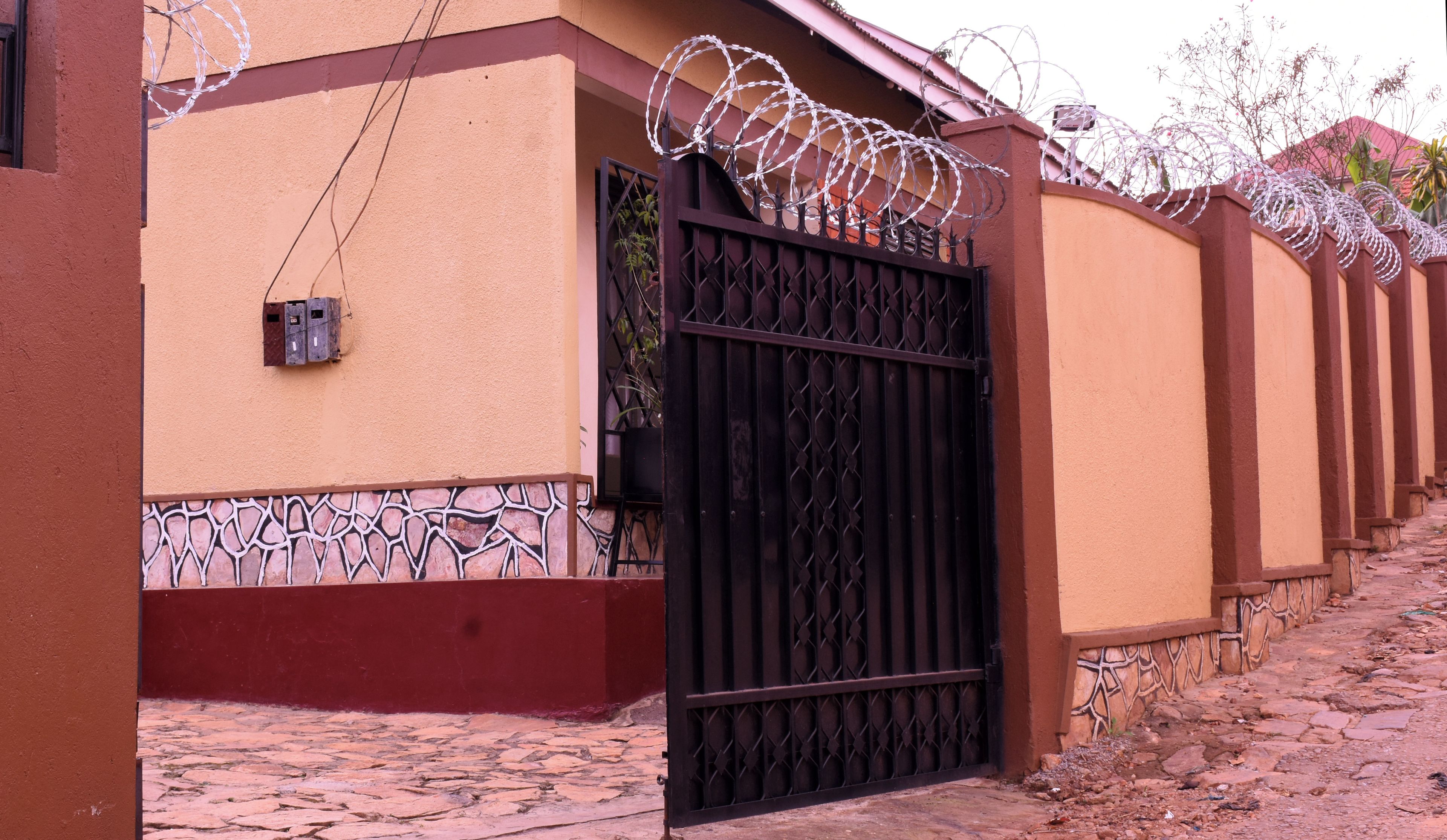 A gated entrance with a metal gate and barbed wire on top, leading to a building with a textured facade and a tiled ground.