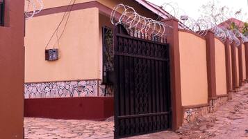 A gated entrance with a metal gate and barbed wire on top, leading to a building with a textured facade and a tiled ground.
