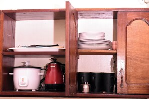 Un mobiletto in legno con ripiani che contiene stoviglie da cucina, tra cui una pentola a cottura lenta bianca, un bollitore elettrico rosso, piatti impilati e tazze nere.