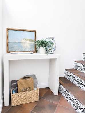 Een witte console tafel met een lijstwerk schilderij, een vaas en een rieten mandje met de tekst 'BEACH' op een trap met decoratieve treden.
