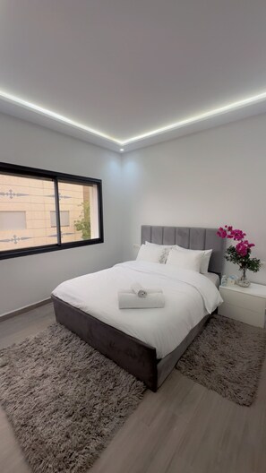 Quarto moderno com uma cama grande, mesa de cabeceira, um vaso com flores e uma janela com vista para edifícios.