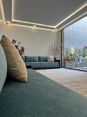 Uma sala de estar moderna com sofá, almofadas e vista para um jardim através de grandes janelas.