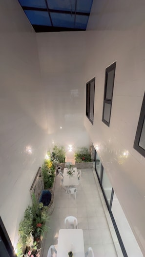 Um alpendre moderno ao ar livre com uma mesa e cadeiras brancas, rodeado por plantas e uma pequena área de jardim.
