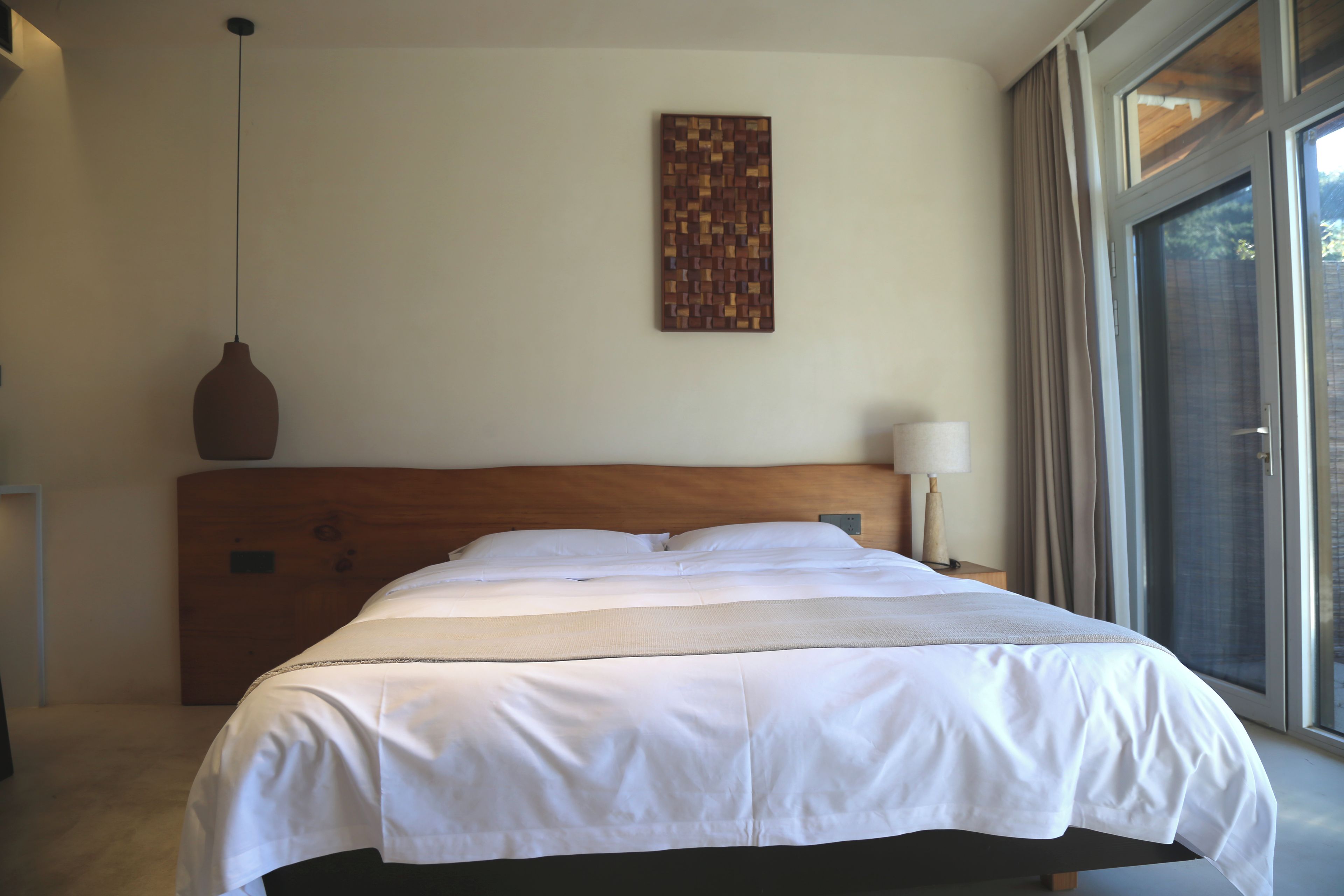 Habitación de hotel con una cama grande, cabecera de madera, una obra de arte colgada en la pared, una lámpara de noche y vistas a través de un cristal.