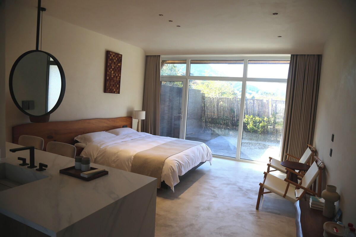 Quarto de hotel com uma cama grande, uma escrivaninha com espelho, uma cadeira e vista para o exterior por uma porta de vidro deslizante.