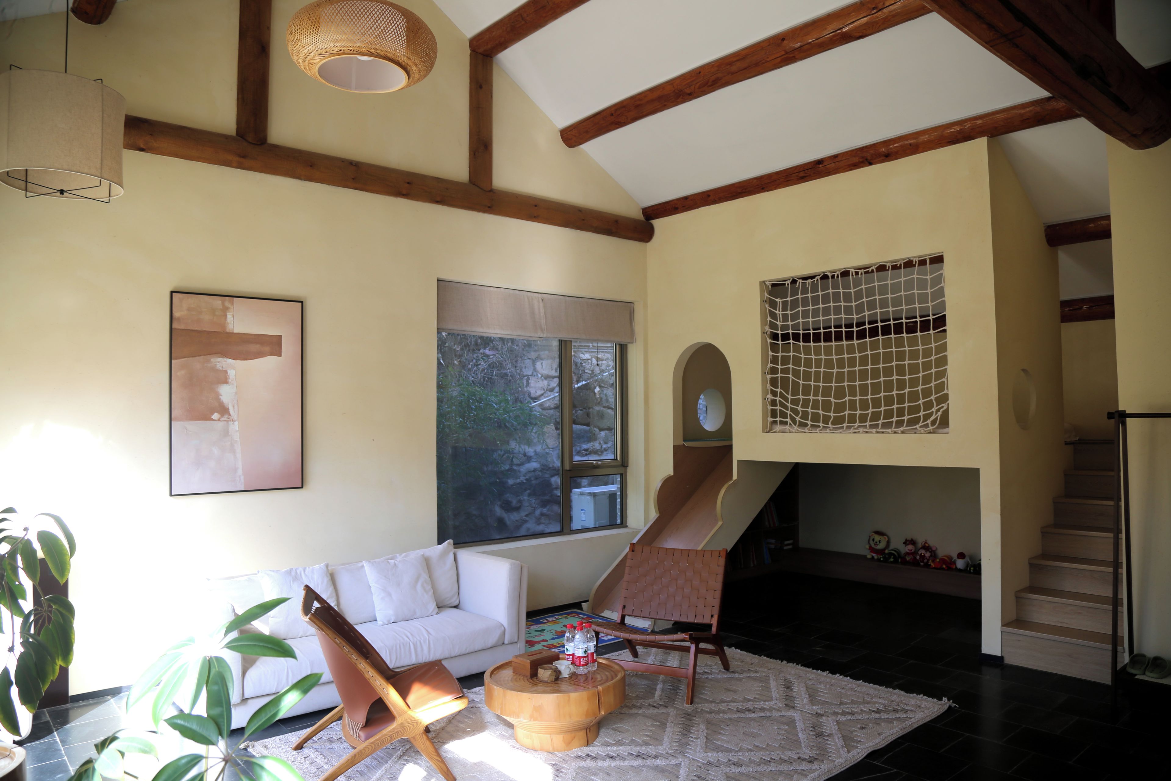 Una sala de estar con un sofá, una mesa de centro de madera, una silla, un cuadro, un ventanal y una escalera.