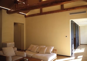 Quarto com sofá, mesa com xícaras, vigas de madeira no teto e porta que dá para outro cômodo.
