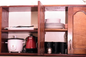 Un mobiletto in legno con ripiani che contiene stoviglie da cucina, tra cui una pentola a cottura lenta bianca, un bollitore elettrico rosso, piatti impilati e tazze nere.