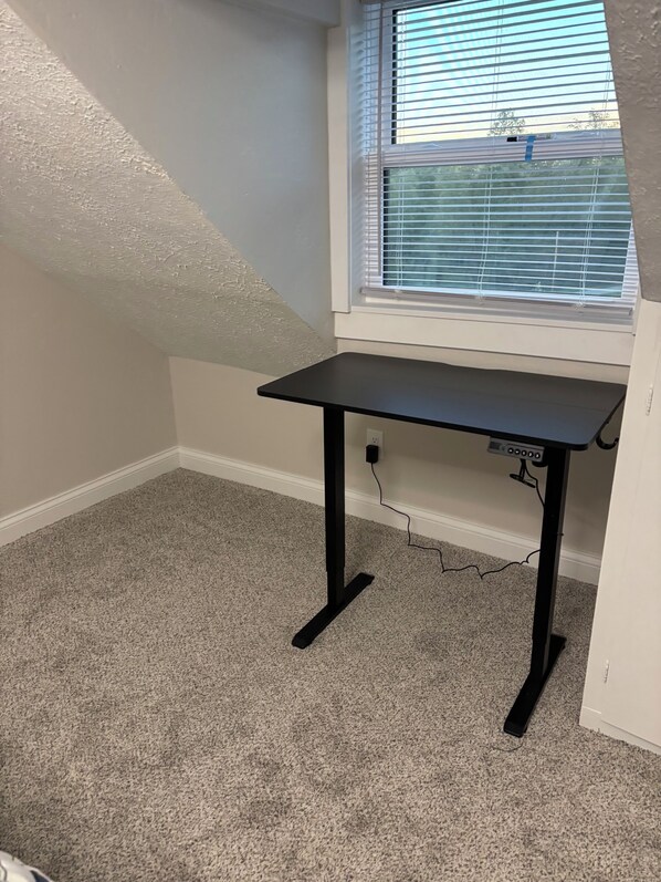 A room with a corner desk, carpeted floor, and a window with blinds.