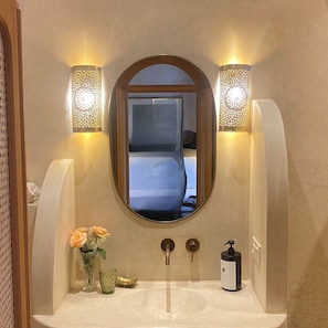 Ein Badezimmer mit Spiegel, zwei Wandleuchten, einem Waschbecken, einer Seifelflasche und einer Vase mit Blumen.