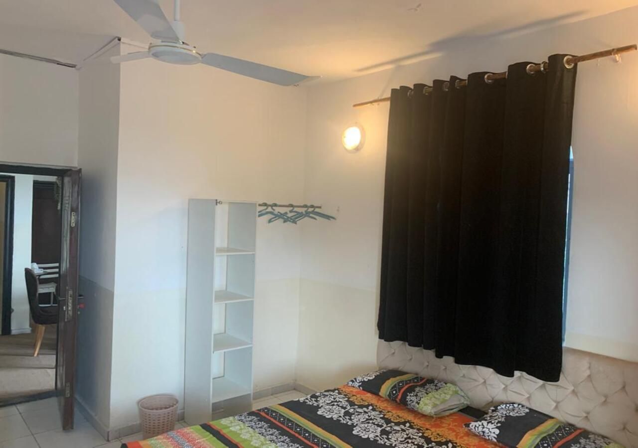 Quarto com cama, ventoinha no teto, guarda-roupa, mesa pequena e janela com cortinas.