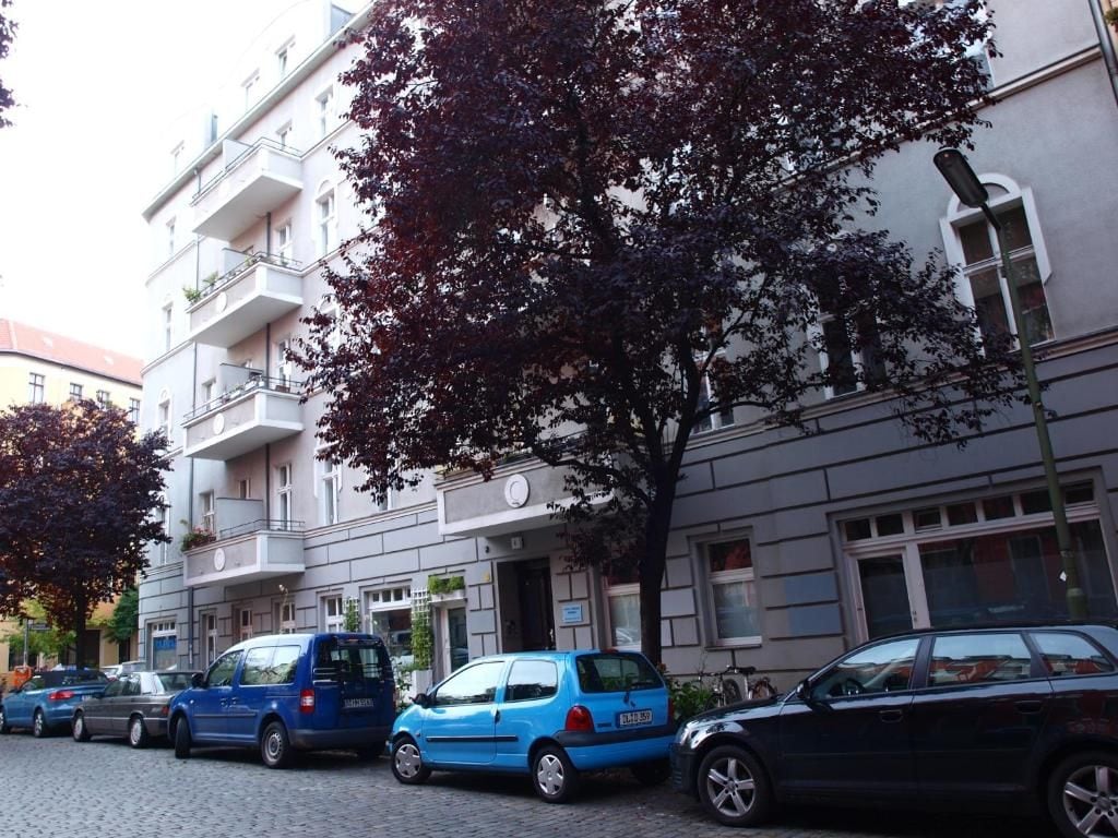 Eine Reihe geparkter Autos säumen eine gepflasterte Straße, im Hintergrund sind Gebäude mit Balkonen und Fenstern zu sehen.