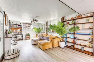 Amplia sala de estar con un sofá seccional grande, una mesa de centro y una estantería llena de libros y objetos decorativos. Hay varias plantas en macetas y un ventilador de techo.