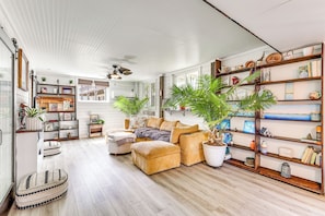 Amplia sala de estar con un sofá seccional grande, una mesa de centro y una estantería llena de libros y objetos decorativos. Hay varias plantas en macetas y un ventilador de techo.