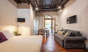 Quarto de hotel com cama, sofá, televisão, mesa de jantar e cadeiras, além de vista pela janela.