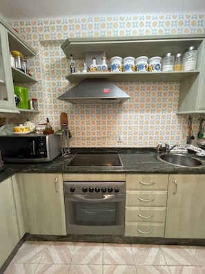 Een keuken met een magnetron, fornuis, oven en een spoelbak. Kastjes en een plank met potten zijn aanwezig.