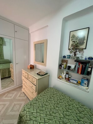 Un dormitorio con un armario, una mesita de noche, una cama con colcha verde a cuadros, un espejo, una estantería con libros y artículos de tocador, y un cuadro en la pared.